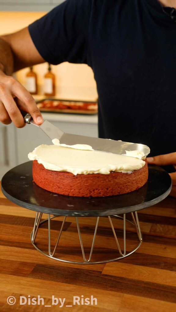 spreading cream cheese frosting over a red velvet cake layer with a spatula