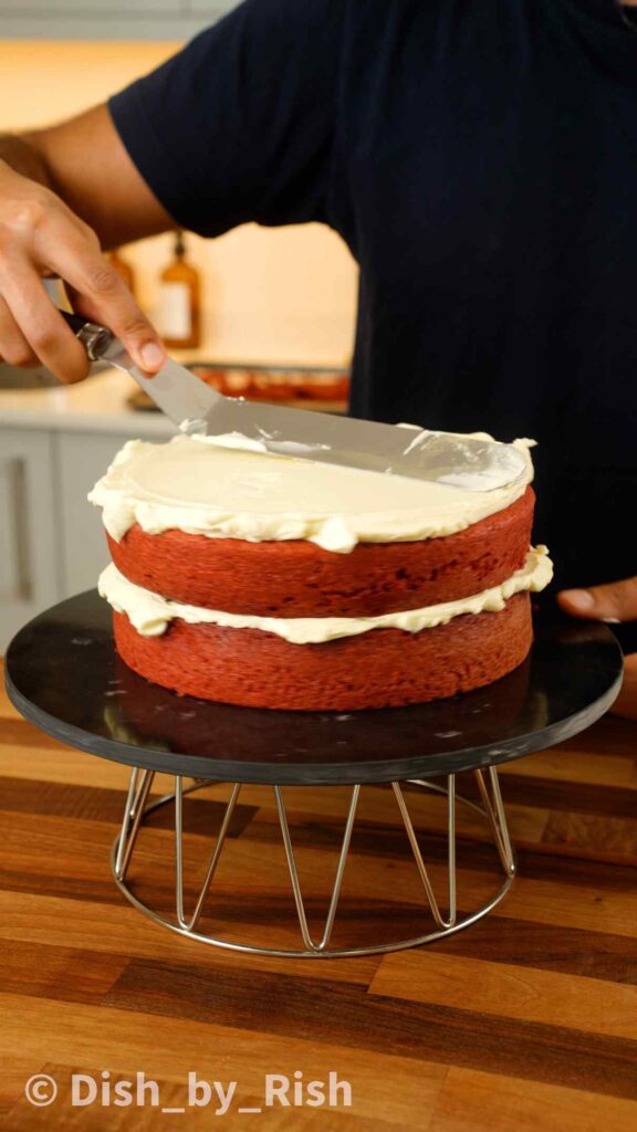 spreading cream cheese frosting over second red velvet cake layer with a spatula