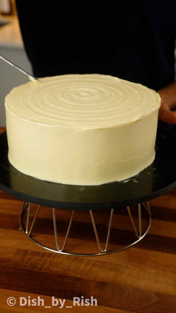 spreading cream cheese frosting all over red velvet cake with a spatula